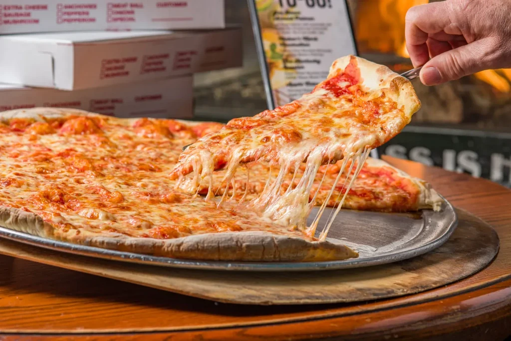 Hand lifts a slice of cooked cheese pizza, cheese stretching.