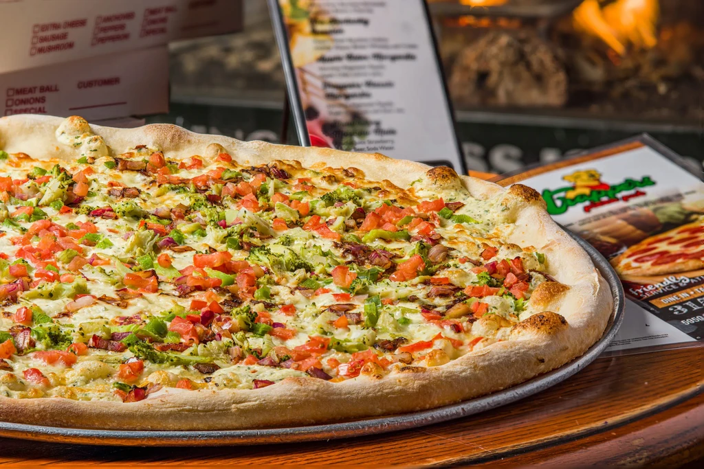 Large rectangular pizza with white sauce, tomatoes, broccoli, bacon in restaurant.