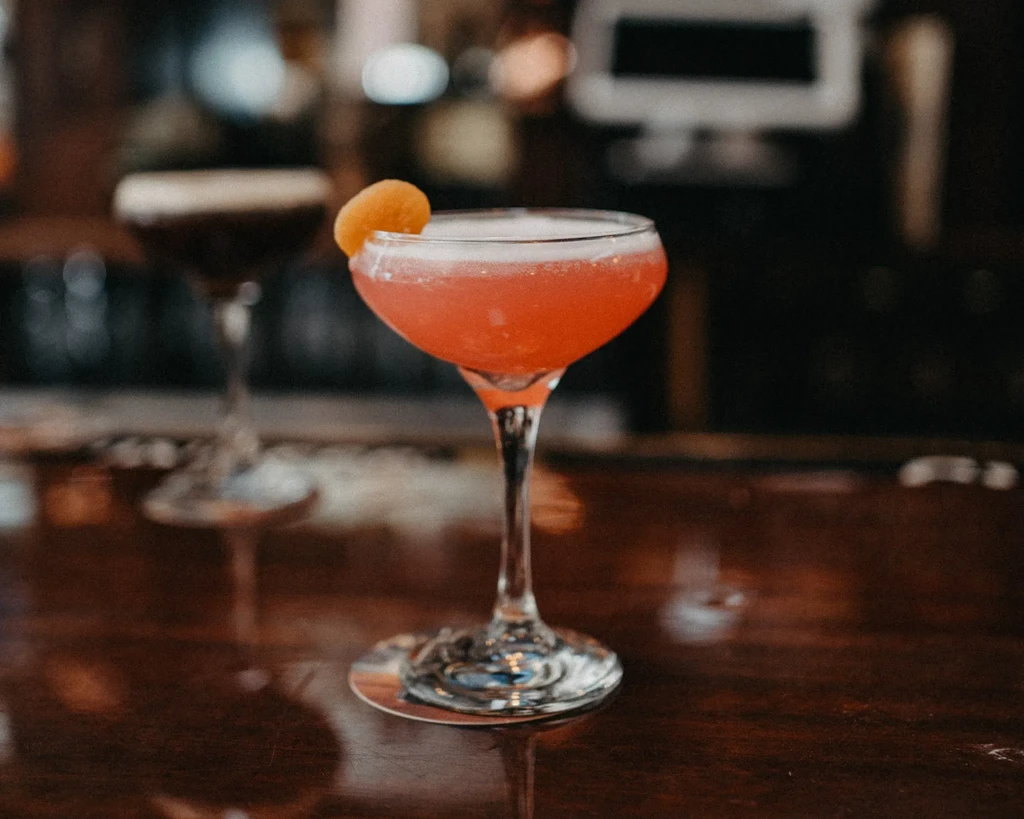 A pink cocktail with orange peel on a wooden bar.