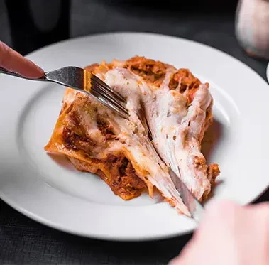 Hands cutting a slice of lasagna with melted, stretchy cheese.