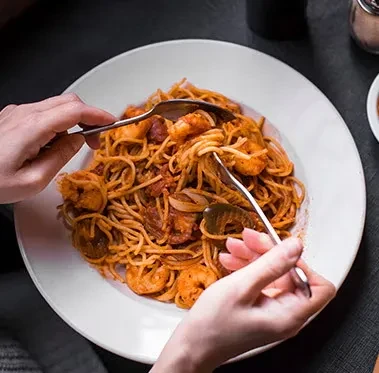 Hands twirling spaghetti with shrimp, meat, and herbs on a white plate.