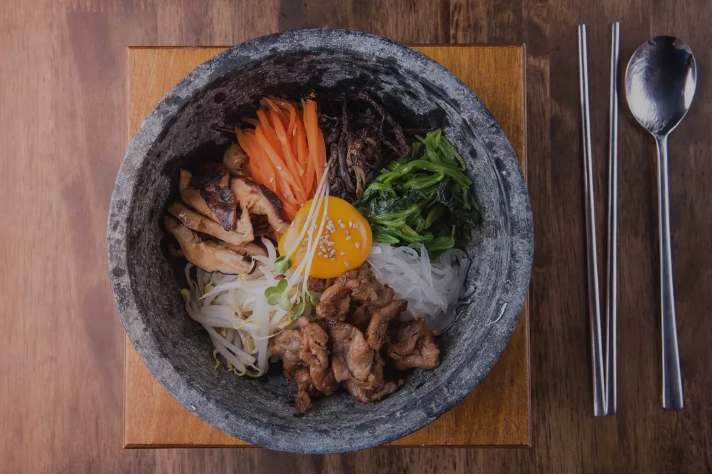 Korean bibimbap in stone bowl with egg, vegetables, meat, chopsticks, spoon.