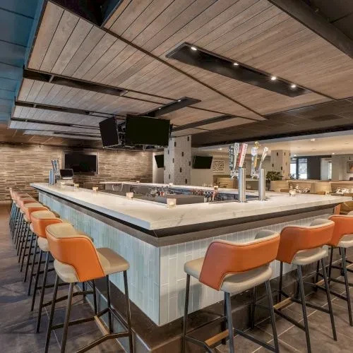 Long, modern bar with orange stools, pale gray tiles, and wood ceiling.