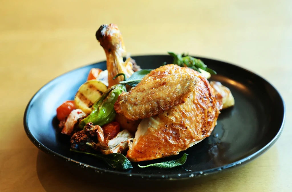 A roasted chicken quarter and drumstick with grilled vegetables on a black plate.