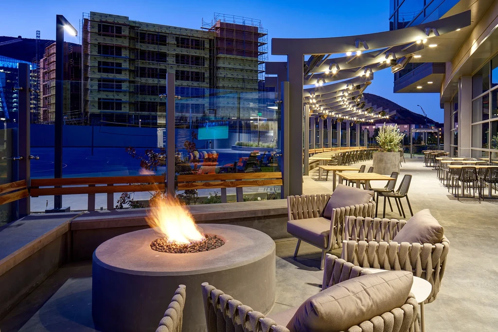 Outdoor patio with fire pit, seating, tables, and city backdrop at dusk.