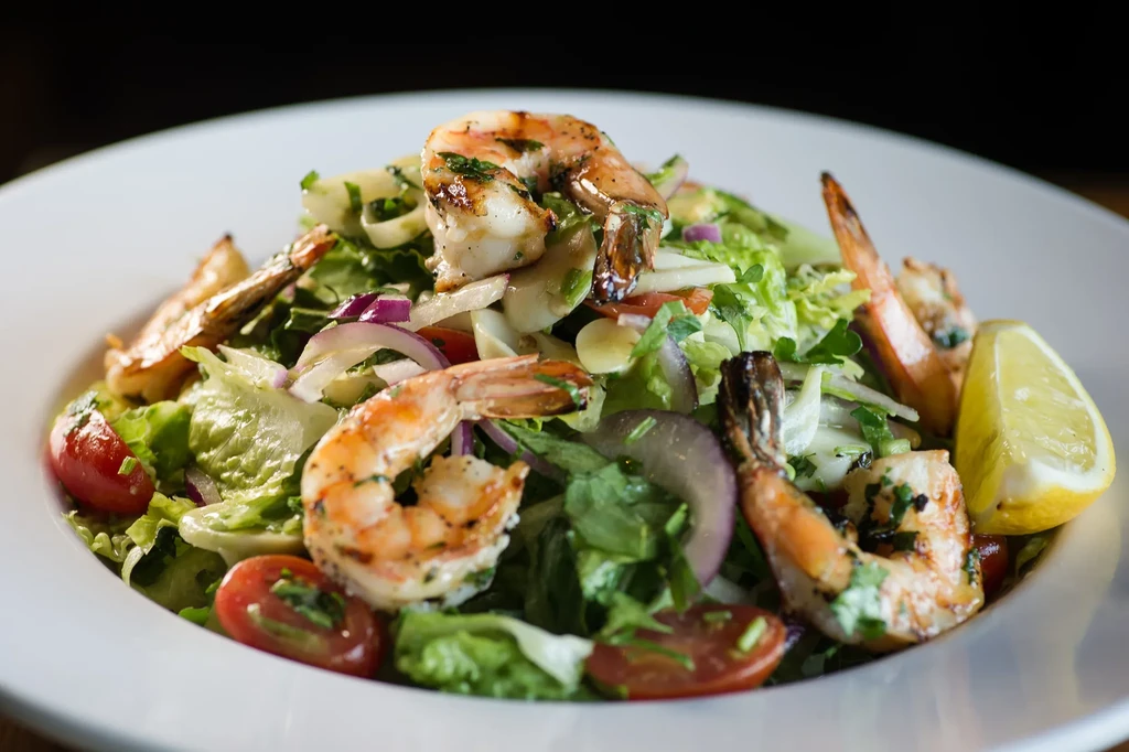 A white bowl filled with grilled shrimp, salad, tomatoes, and lemon.