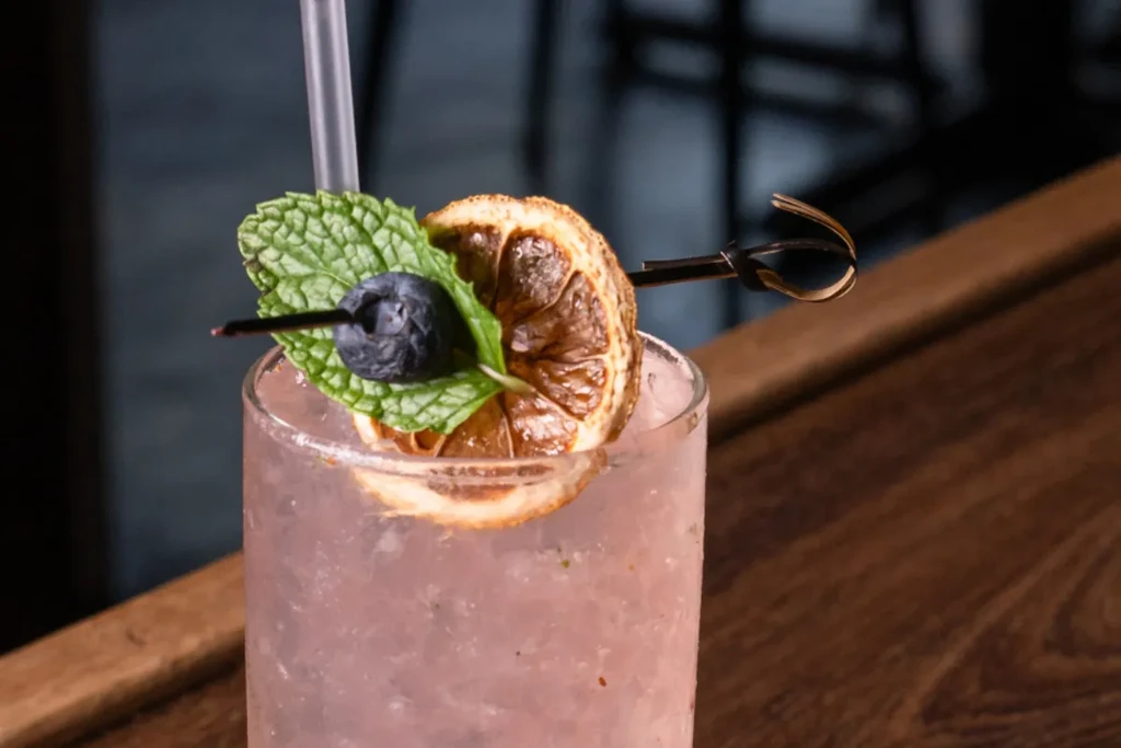A purple cocktail garnished with a blueberry, mint leaves, and dried orange.