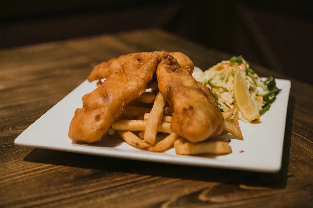 A white plate holds two pieces of fried fish, fries, and coleslaw.