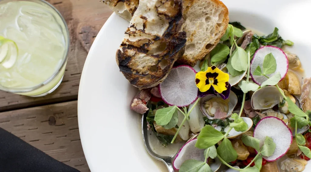 A bowl of clams, radishes, greens, grilled bread, and a drink.