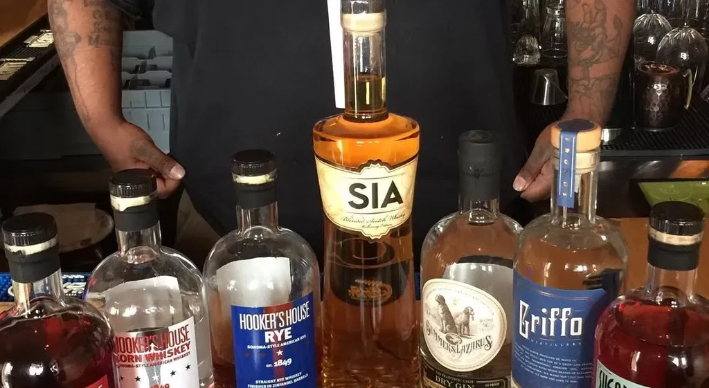 A person with tattooed arms stands behind a collection of liquor bottles.