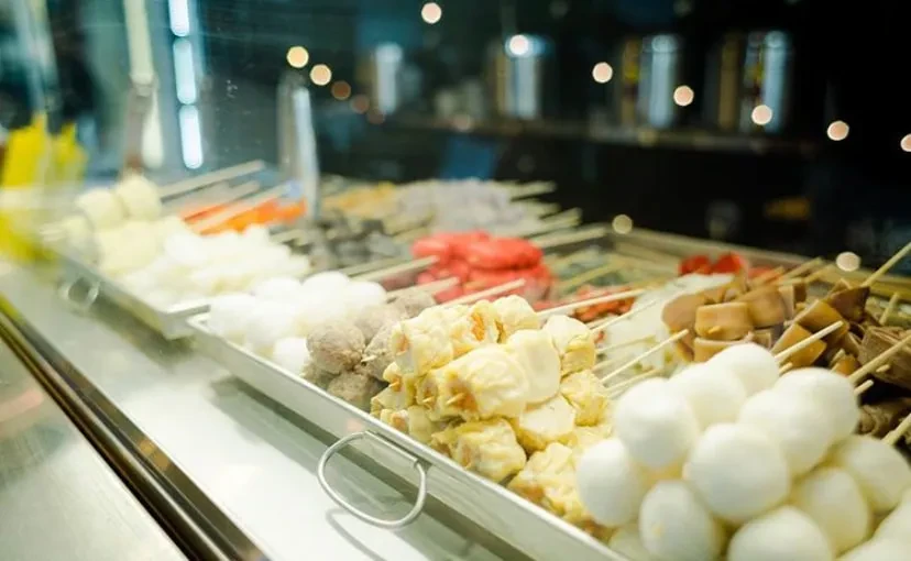 Skewers of street food, including dumplings, meatballs, and tofu, behind glass.