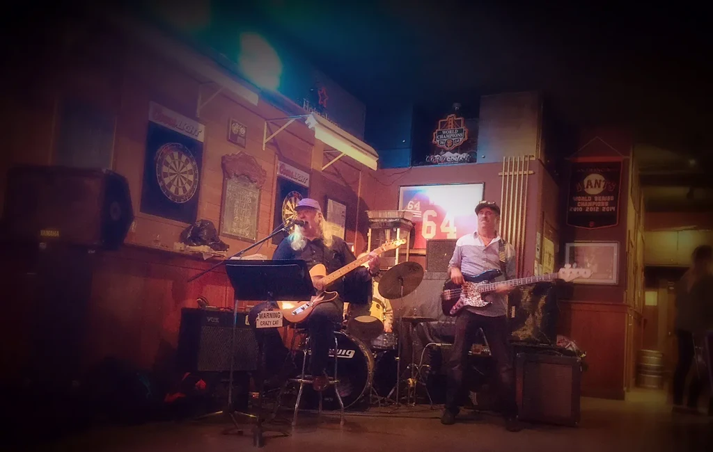 Two men play electric guitar and bass in a dimly lit bar.