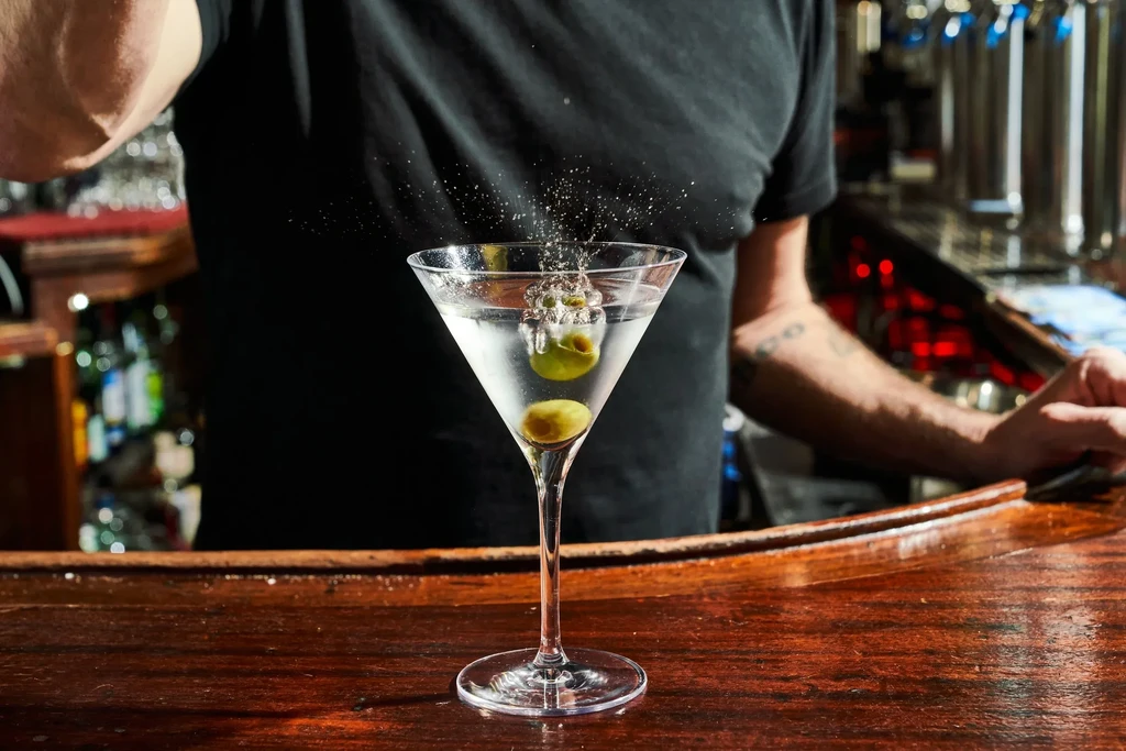 Martini with olives splashing into it on a wooden bar counter.