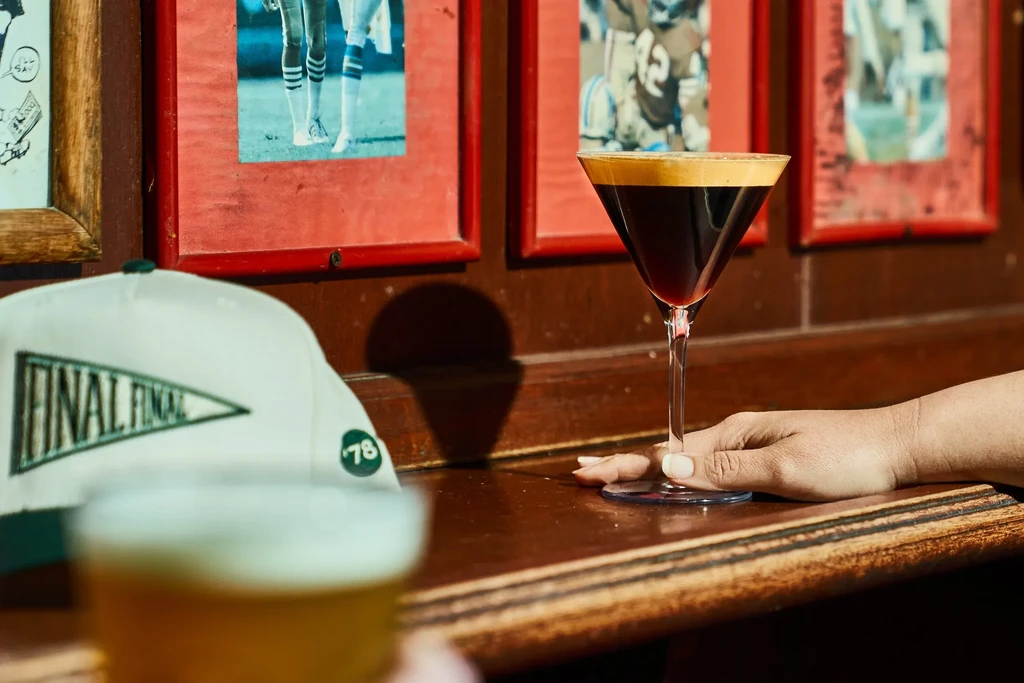 Hand holding a martini glass on a bar next to a white hat.