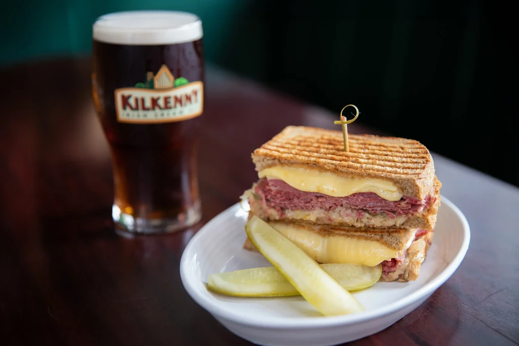 Kilkenny beer, a Reuben sandwich, and pickle spears on a dark table.