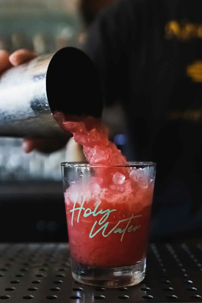 Bartender pours red drink with ice into a "Holy Water" glass.