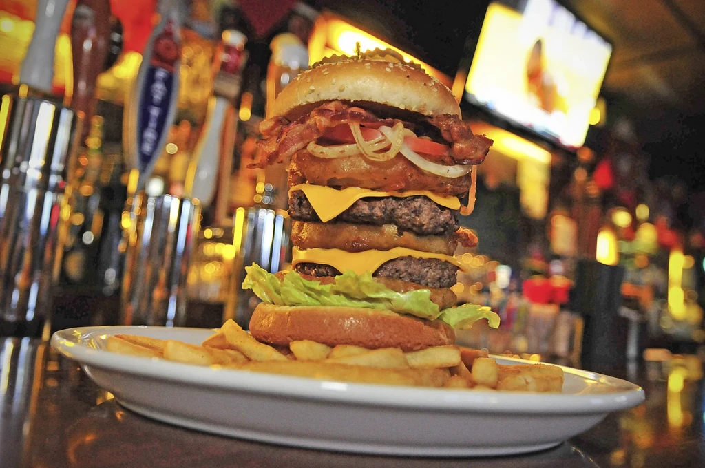 A massive, stacked hamburger with fries on a plate in a dimly lit bar.