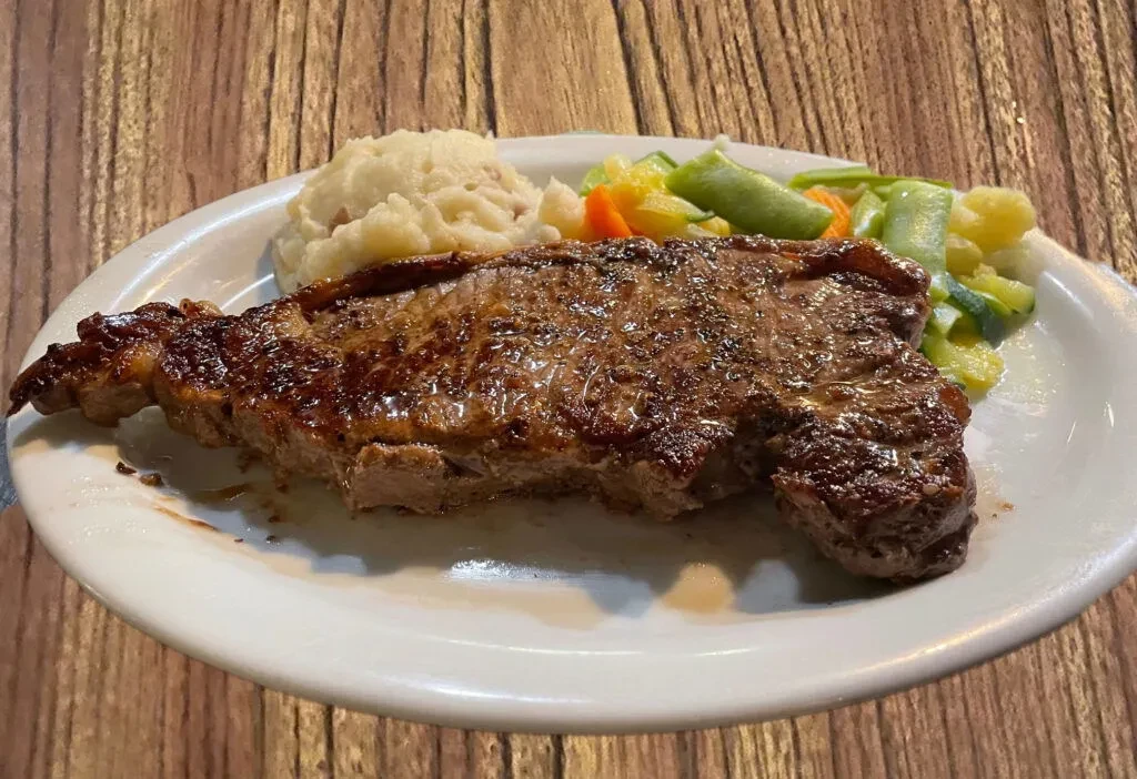 A large steak, mashed potatoes, and mixed vegetables on a white plate.