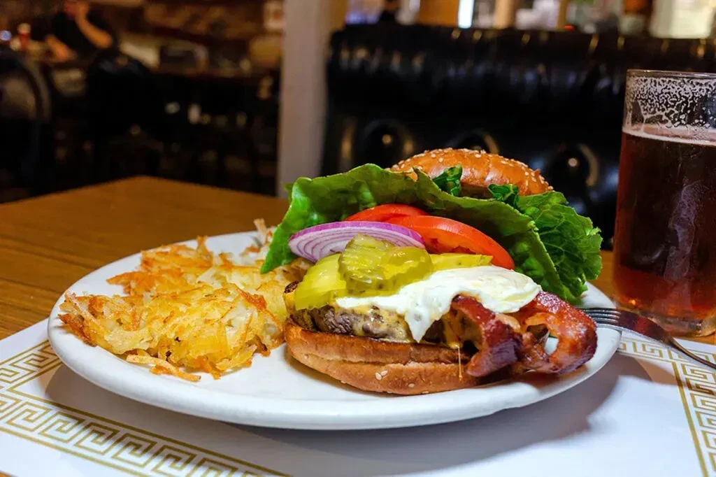 A bacon cheeseburger with hash browns and a drink on a white plate.