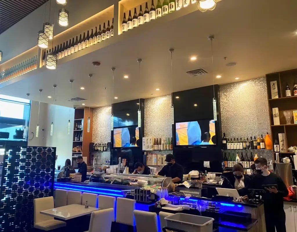 Modern Asian restaurant with a sushi bar, shelves of bottles, and servers.