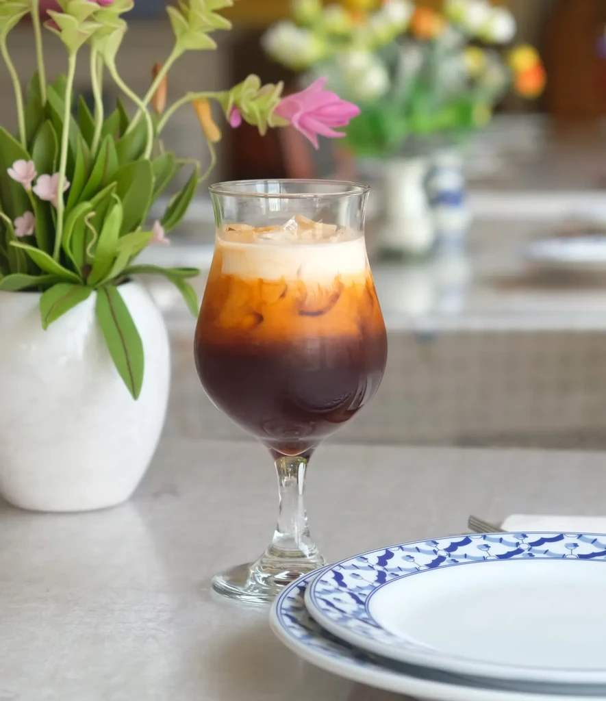 A layered iced tea drink in a wine glass beside a plant and plates.
