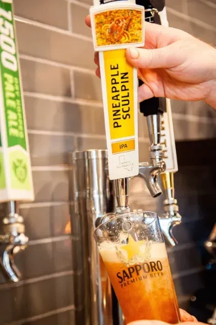 A person pours a glass of Pineapple Sculpin IPA from a beer tap.