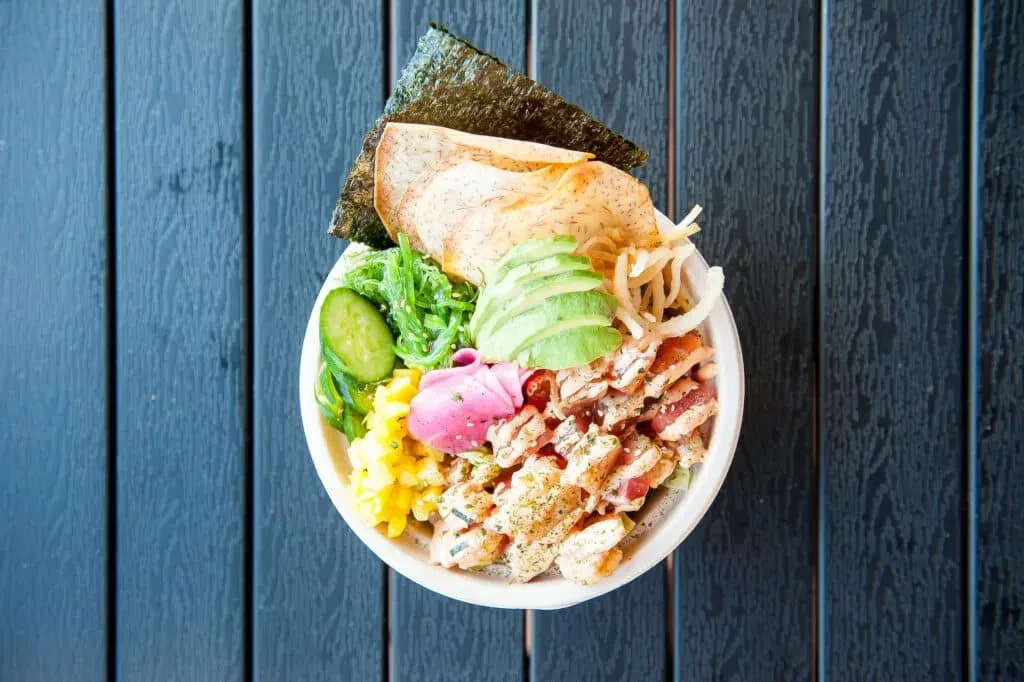Top-down view of poke bowl with avocado and seaweed on dark wood.