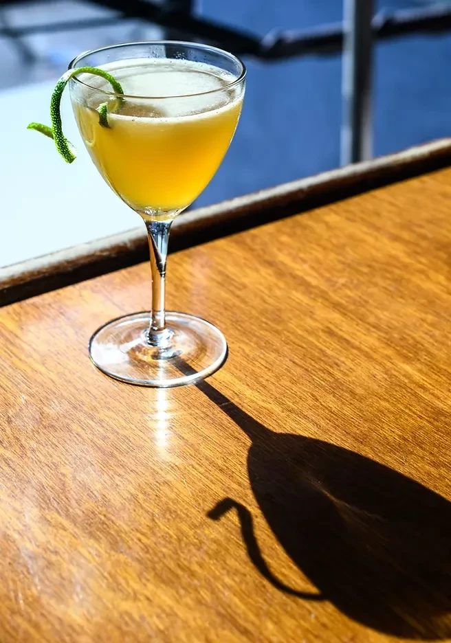 A yellow cocktail with a lime peel garnish on a wooden table.