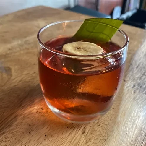A glass of amber liquid garnished with a banana slice and leaf.