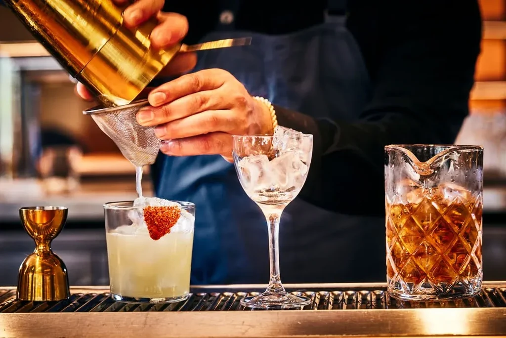 Bartender straining a cocktail into a glass on a bar counter.