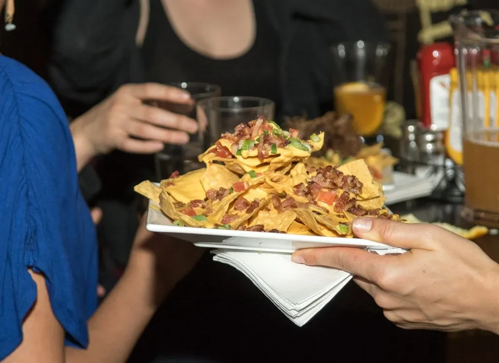 A hand holds a plate of loaded nachos with bacon and tomatoes.