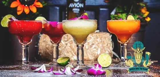 Four colorful margaritas with lime wedges and flower petals on a table.