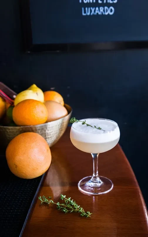 A light-colored cocktail with foam and a thyme sprig.