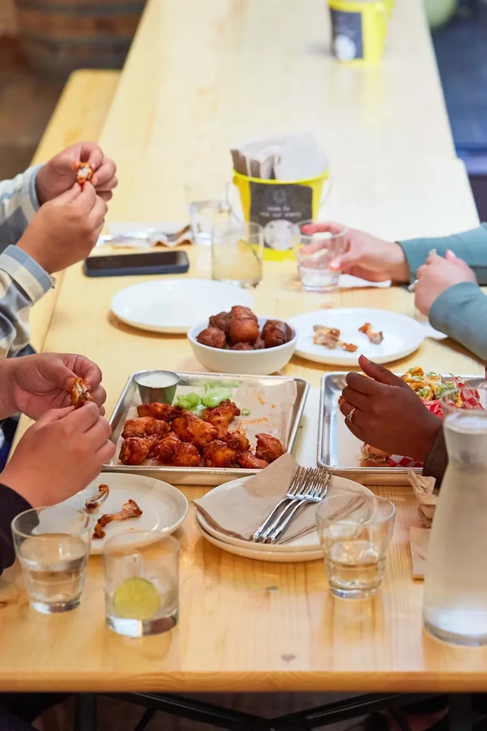 People eating wings and fried food at a long wooden table.