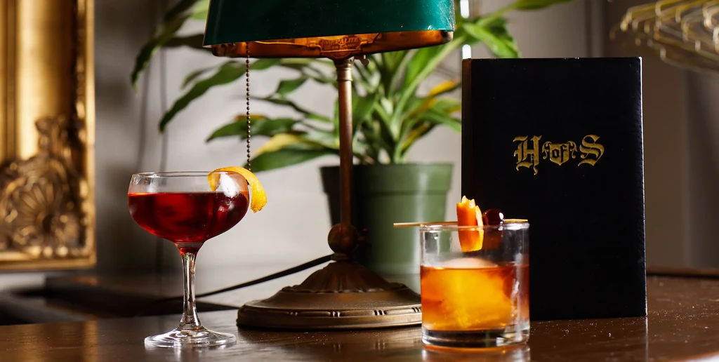 Two cocktails on a table with an antique lamp and a menu.
