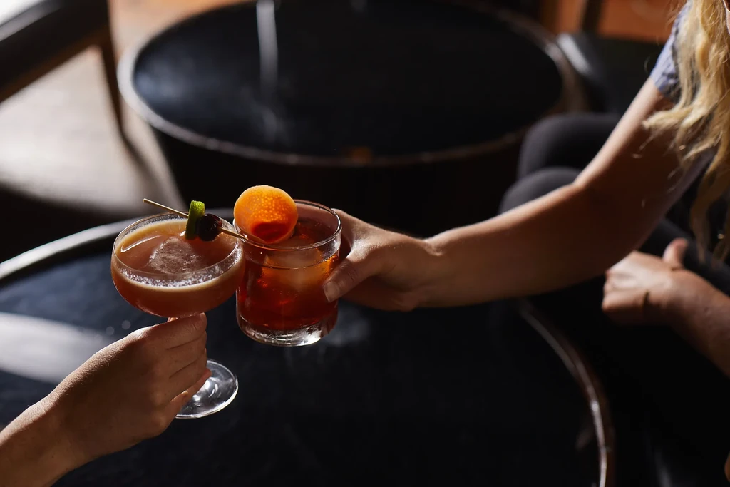 Two hands clinking cocktails at a dimly lit bar.