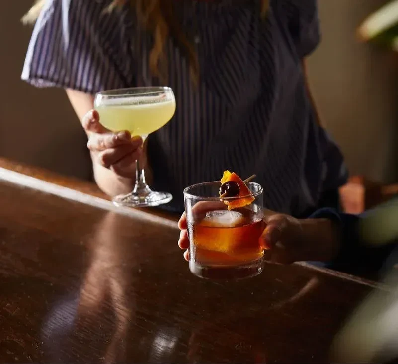 Two cocktails, one yellow, one amber, held by hands at a bar.