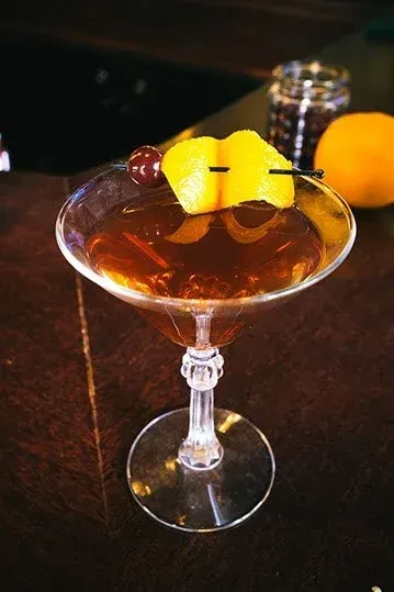 A brown cocktail in a coupe glass with a cherry and lemon twist.