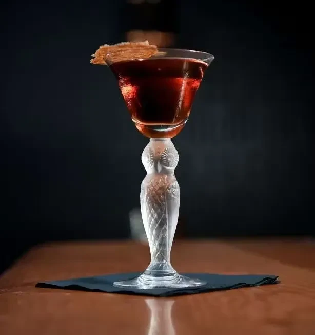 A dark red cocktail in a clear owl-stemmed glass with a cracker garnish.