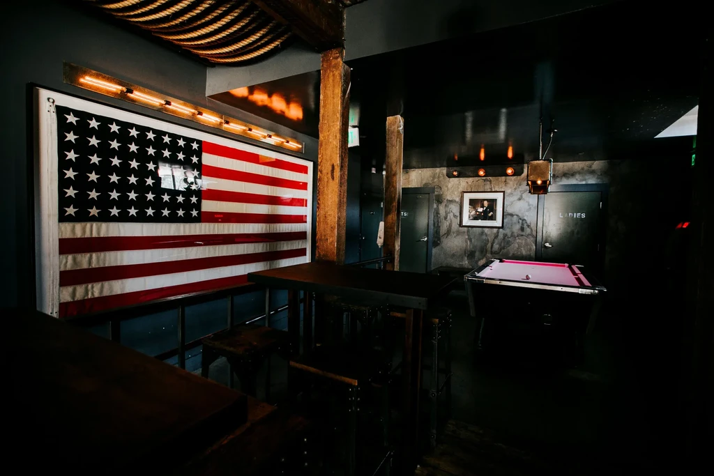 Dimly lit bar with an American flag, dark tables, and a pink pool table.