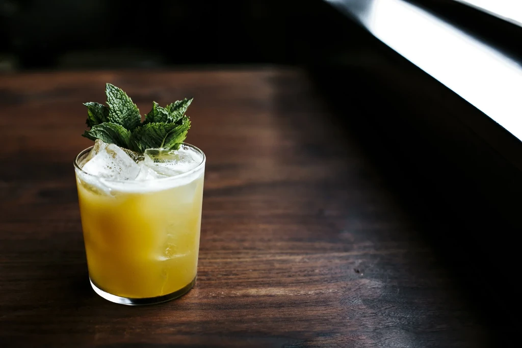 A glass of iced yellow cocktail with mint leaves on a dark wooden table.