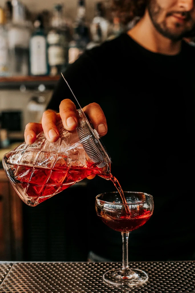Bartender pours a red drink from a mixing glass into a stemmed cocktail glass.