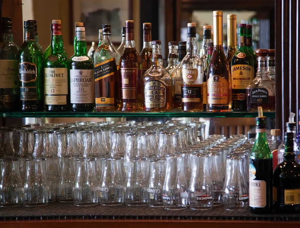 Various liquor bottles sit on a glass shelf above rows of empty glasses.