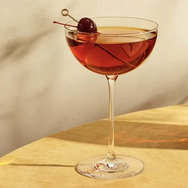 Coup glass of red cocktail with cherry garnish on yellow table.