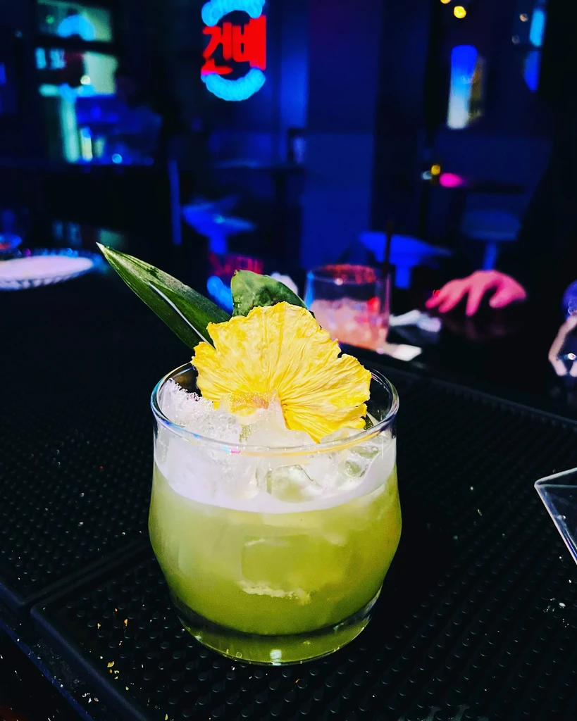 A green cocktail with a dried pineapple and leaf garnish on a bar.