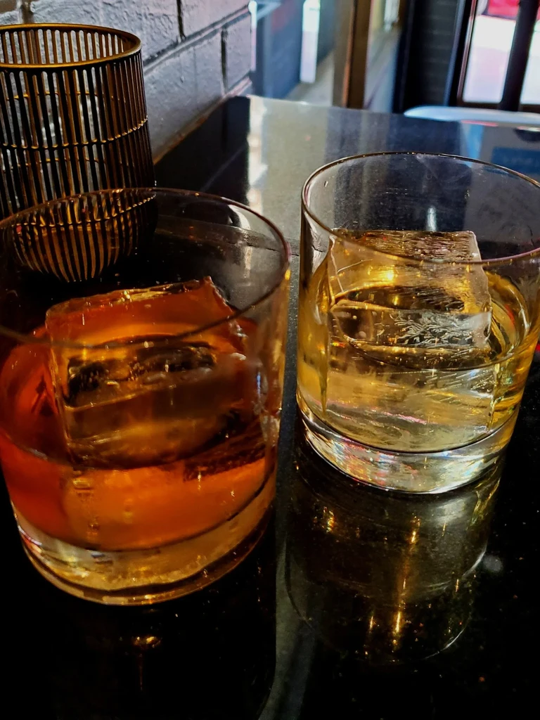 Two glasses of whiskey with ice on a reflective black surface.