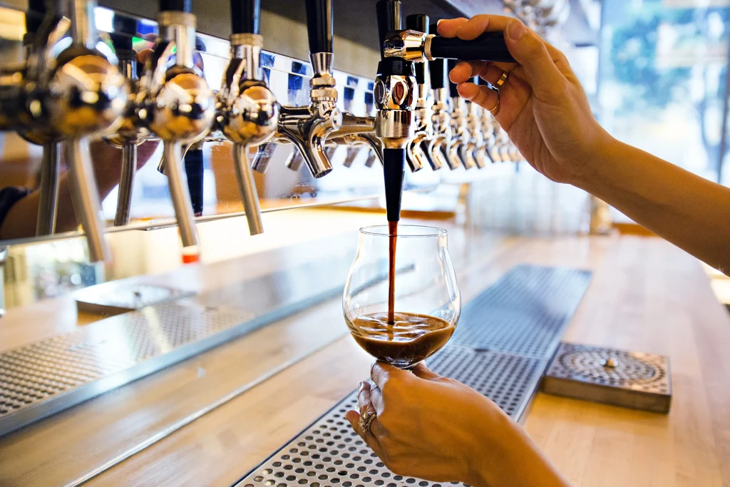 A person pours dark liquid from a tap into a stemmed glass.