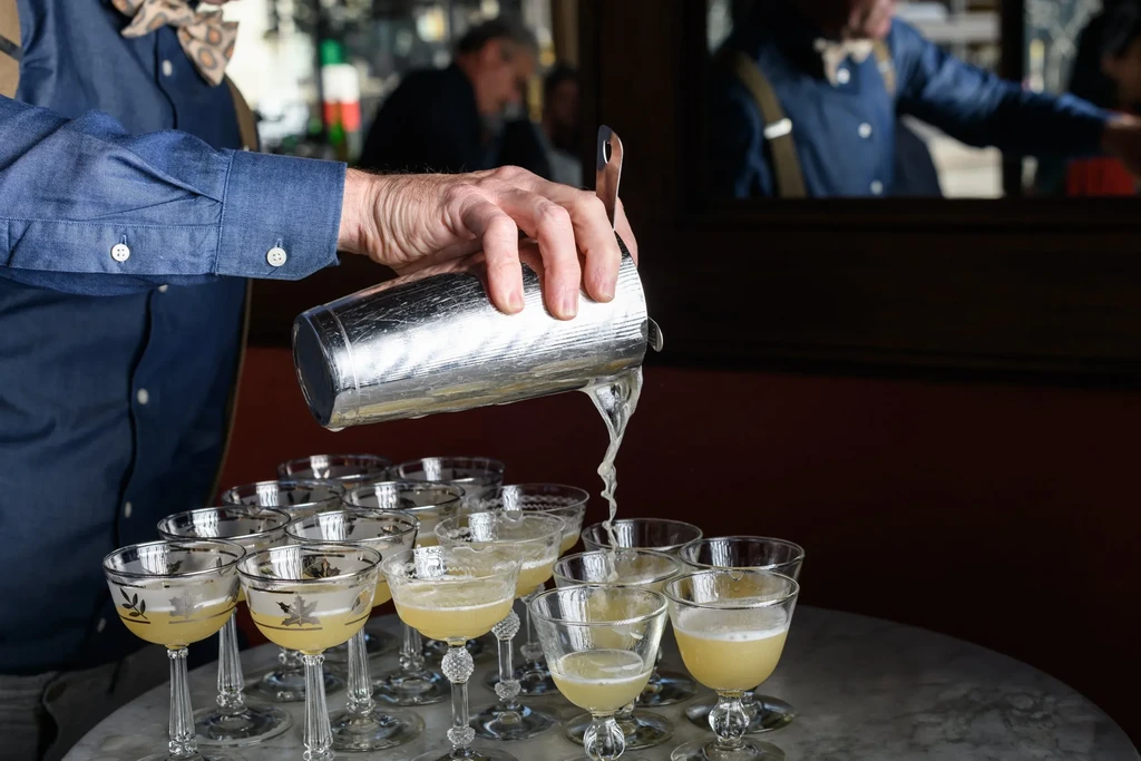 A bartender pours light yellow liquid from a shaker into multiple glasses.