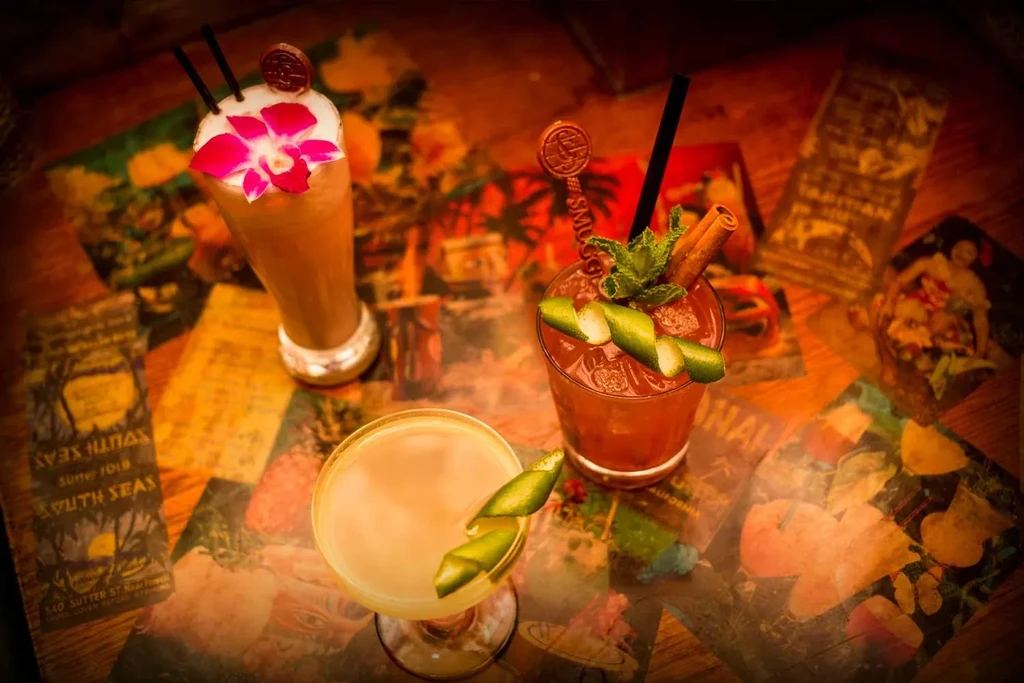 Three cocktails with garnishes sit on a colorful, patterned table.
