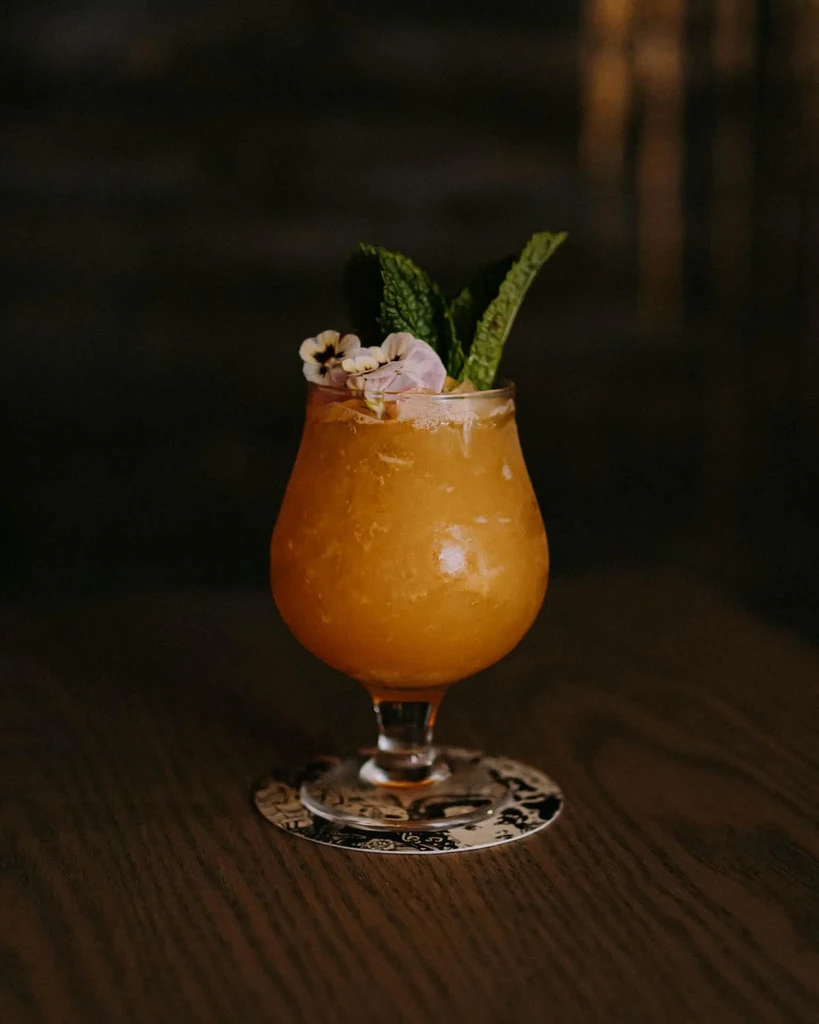 An orange cocktail in a stem glass garnished with mint and flowers.